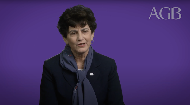 Still image from a video showing a woman wearing a dark suit and purple scarf in front of a purple background that includes the letters A, G, B.