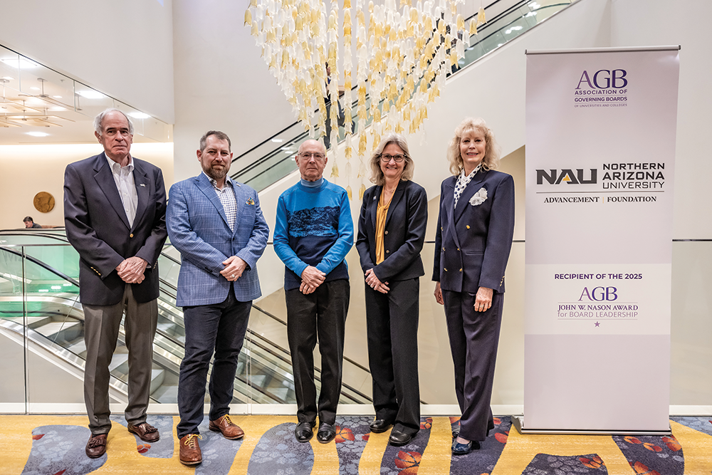Five individuals stand in a row at an award ceremony for the Northern Arizona University Foundation Board. From left to right: William Heibel in a dark suit with light-colored shirt, Nick Dolce (associate vice president of college fundraising) in a blue checkered blazer, Robert Braudy wearing a blue turtleneck sweater, Victoria Fimea (board chair) in a black blazer with yellow blouse, and Lisa Bagwell (vice chair) in a navy suit with patterned scarf. They are positioned in front of a modern interior with an escalator and decorative hanging installation visible in the background. To the right is a banner displaying the NAU (Northern Arizona University) Advancement Foundation logo and text reading 'RECIPIENT OF THE 2025 AGB JOHN W. NASON AWARD for BOARD LEADERSHIP.' The AGB (Association of Governing Boards of Universities and Colleges) logo appears at the top of the banner.