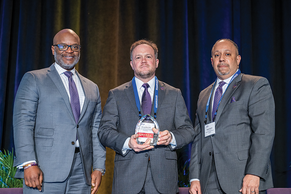 Three men in suits stand together at an award ceremony. From left to right: Alfonzo Alexander (board chair) wearing a light gray suit with purple tie and glasses, Kevin Braswell (executive director) in a darker gray suit holding a crystal award labeled 'Speaker's Forum Honorable' with a blue lanyard around his neck, and Nathan Burton (vice chair-elect) in a gray suit with a blue lanyard and name badge. The background features blue curtains, and the image shows The Tennessee Technological University Foundation Board accepting the Nason Award. A small plant is partially visible in the lower left corner.