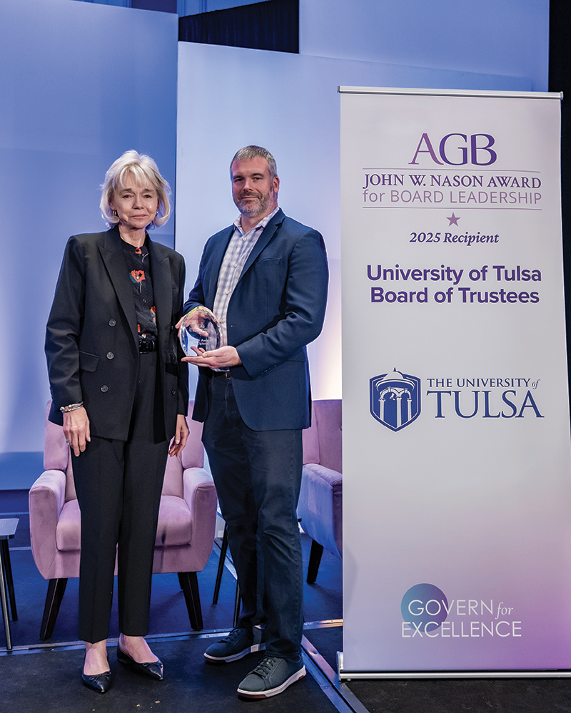 Two individuals at an award ceremony standing side by side. On the left is Marcia MacLeod (University of Tulsa Board Chair) wearing a black suit with a patterned top underneath. On the right is Robert Thomas (Finance Committee Chair) in a blue suit holding a crystal award. They're standing in front of a banner displaying 'AGB JOHN W. NASON AWARD for BOARD LEADERSHIP' with '2025 Recipient University of Tulsa Board of Trustees' and the University of Tulsa logo below. The background features blue lighting and there's a light-colored chair visible. The bottom of the banner shows 'GOVERN for EXCELLENCE.